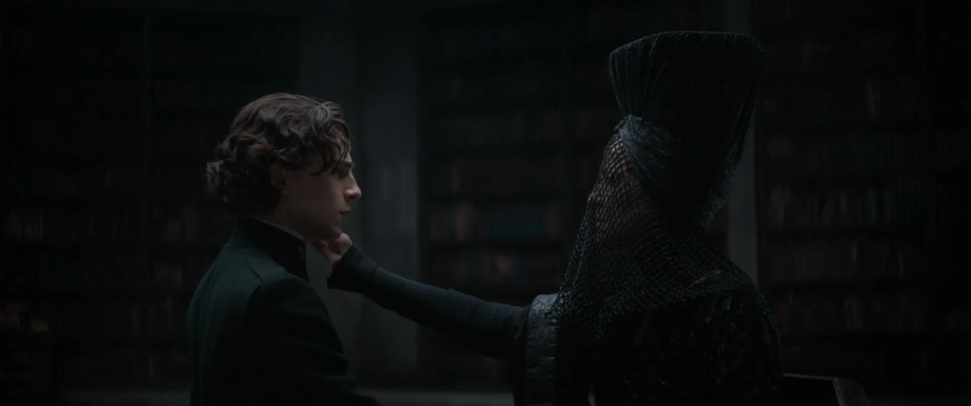 A young man in black extends his hand toward a hooded figure in dark armor, in a dim library-like room