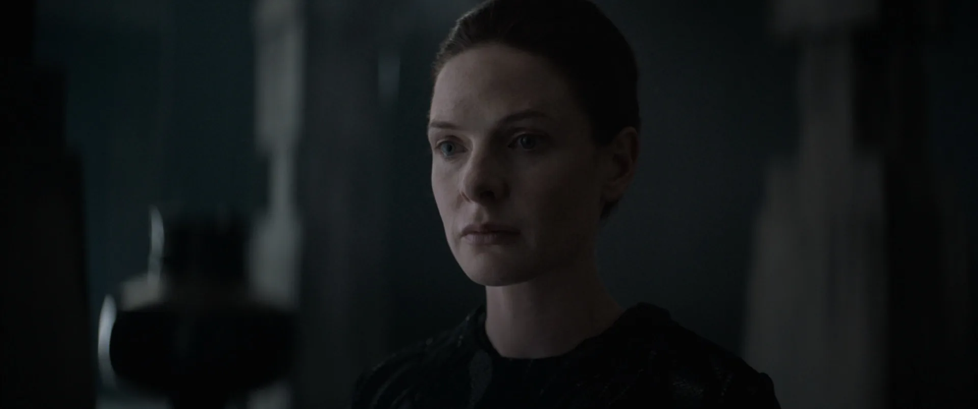 Portrait of a woman with tied-back hair, dressed in black, with a fixed gaze and grave expression, in a dim setting