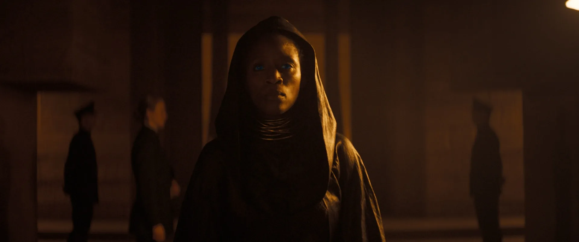 Figure wearing a dark hood and a metallic collar, with intensely blue eyes, standing in a large amber-lit hall with silhouettes in the background